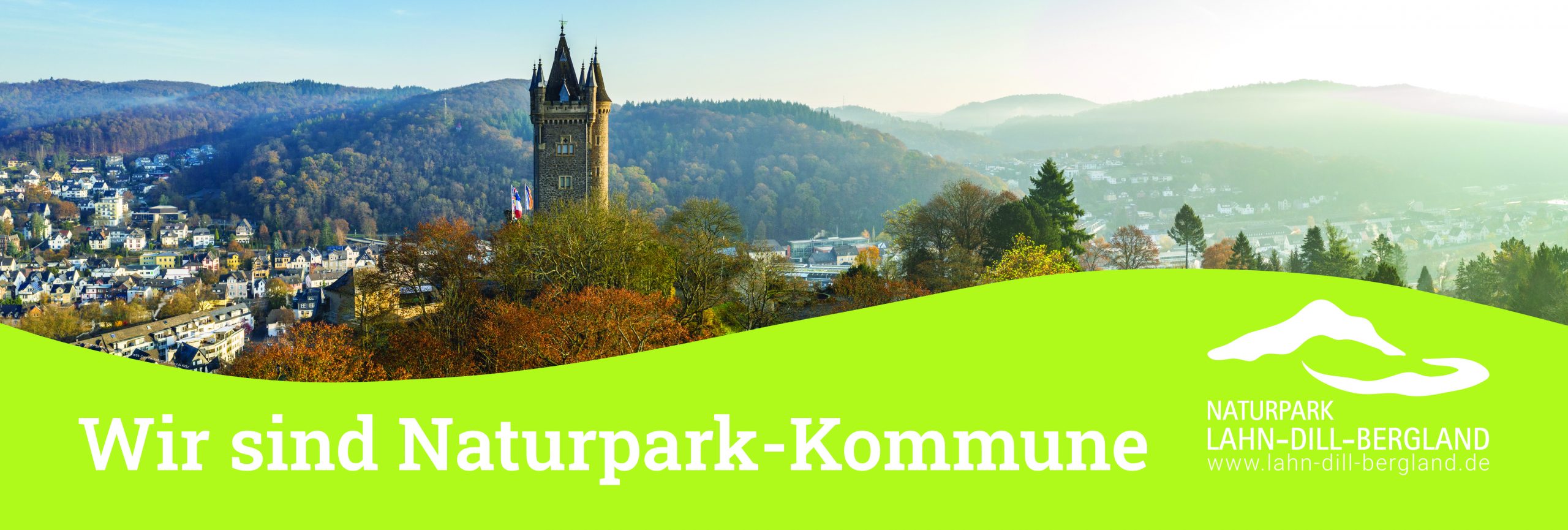 Banner "Wir sind Naturpark-Kommune". Der obere Teil des Banners zeigt eine Stadtansicht von Dillenburg mit dem Wilhelmsturm. Der untere Teil ist ein hügelartiges grünes Feld in dem die Worte "Wir sind Naturpark-Kommune" und das Logo des Naturparks Lahn-Dill-Bergland in weiß stehen.